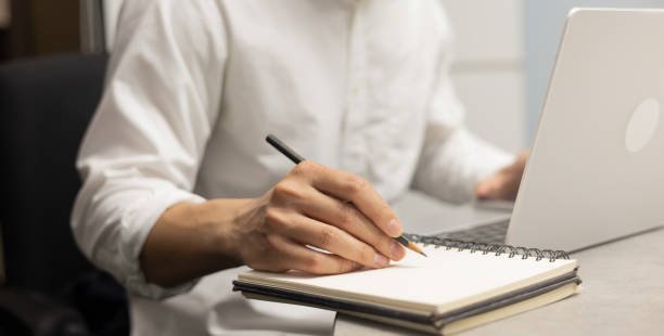 Close-up of a businessman’s hand writing in a notebook while working on a laptop at a wooden desk in a home office.