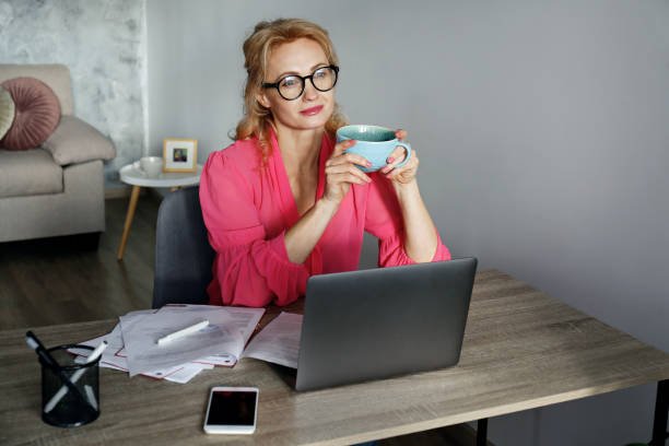 Adult blonde businesswoman sitting at her desk with a laptop, holding a large cup of coffee during a work break
