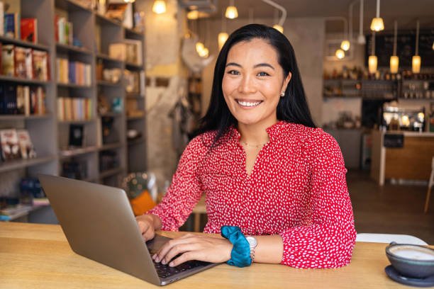 Young Asian woman smiling while e-learning on her laptop in a modern cozy café, wearing earphones and sitting at a wooden table with a coffee cup.