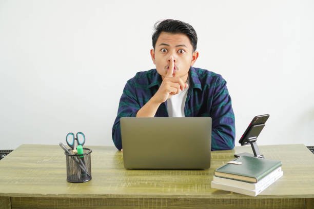 Asian male freelancer sitting at a desk with a laptop, making a silent gesture by placing his finger on his lips.