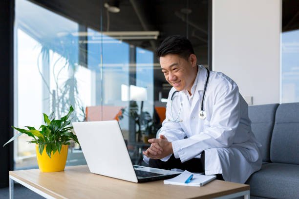Friendly Asian doctor using a laptop for remote consultation with a patient, engaging through webcam and monitor in a clinic.